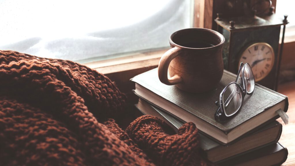 Um canto de janela com um cobertor, uma pilha de livros, uma xícara de café, um relógio em cima deles. Logo ao fundo e um óculos.