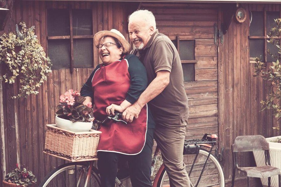 Casal de idosos rindo, andando em uma bicicleta, vivendo a envelhescência com plenitude.