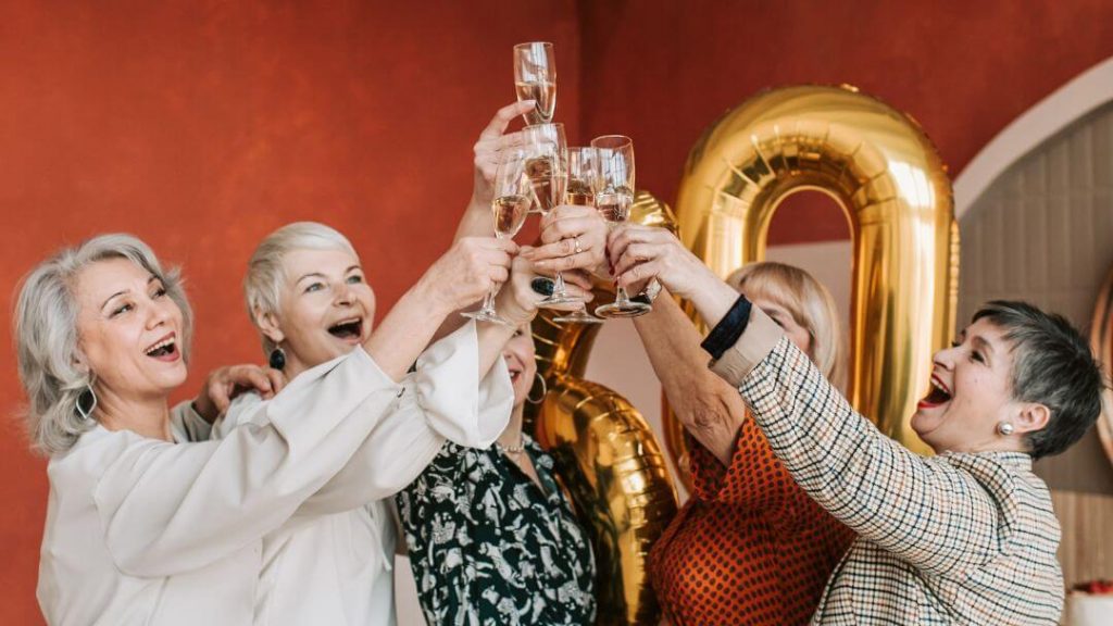 Grupo de cinco mulheres idosas brindando com champanhe o aniversário de uma delas e a envelhescência.