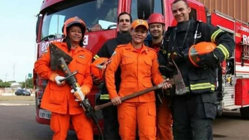 Pequena grupo de funcionários do Corpo de Bombeiros, contendo três homens e duas mulheres, posam para foto vestidos com o uniforme, e segurando alguns instrumentos de trabalho em frente ao caminhão de bombeiros.