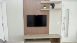 A sala mostra um raque de MDF, com cores claras. O painel onde está pendurada TV Led, tem cor clara e aspecto que imita madeira. Abaixo da TV um aparador na cor bage. Acima da TV, do lado direito, duas prateleiras da mesma cor do aparador.