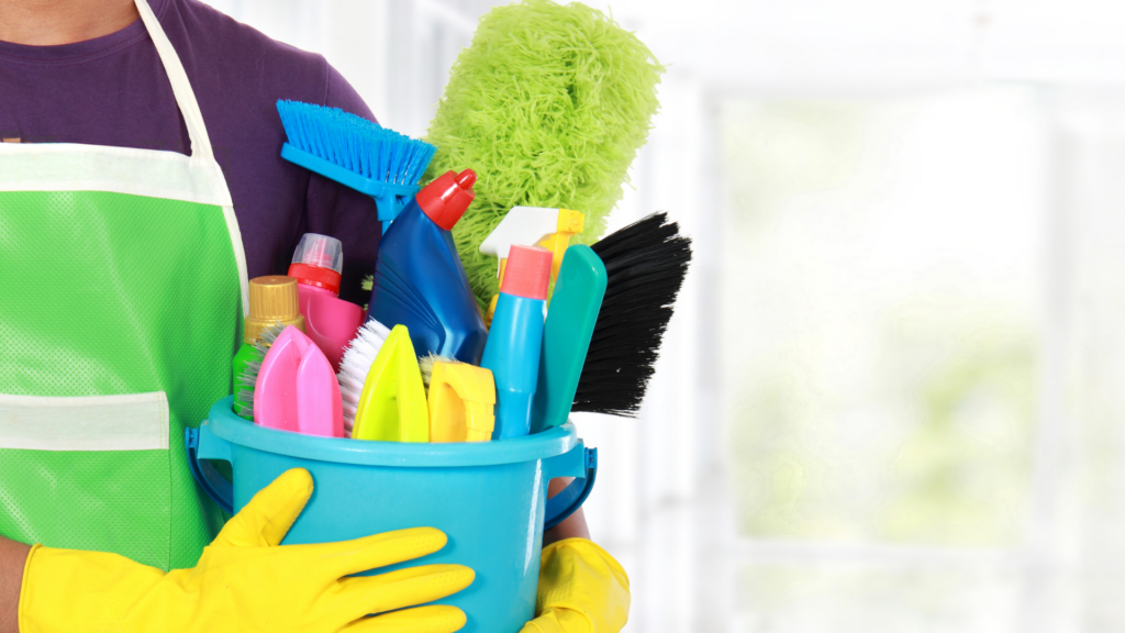Parte de homem jovem carregando um balde com produtos e escovões dentro. Ele usa luvas de limpeza amarelas. Ele está se preparando para limpar a casa.