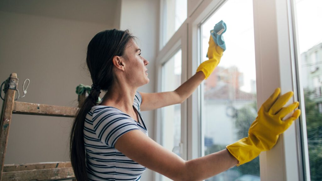 Mulher branca de cabelos pretos longos presos com um rabo baixo. Ela usa luvas e está passando um pano no vidro da janela para limpar a casa.