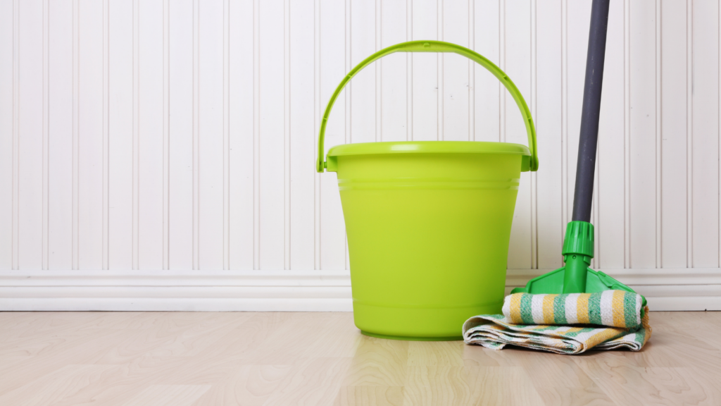 Dica importante é utilizada materiais corretos para limpar a casa.
Um balde verde neon com alça levantada. Ao lado um esfregão de pano, com ponta verde e cabo preto. O pano tem as cores branco, amarelo e verde.
A parede tem revestimento de plástico branco na vertical.