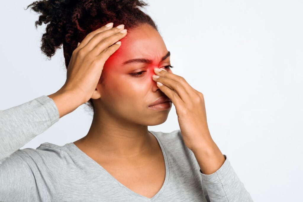 Mulher jovem negra com face de dor e desconforto, colocando uma mão no nariz e outra mão na cabeça. O nariz e a cabeça estão vermelhos.