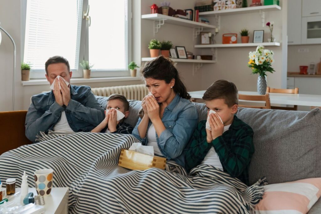 Família composta de pai, mãe e deois filhos crianças, todos sentados no sofá assoando o nariz por estarem gripados ou resfriados.