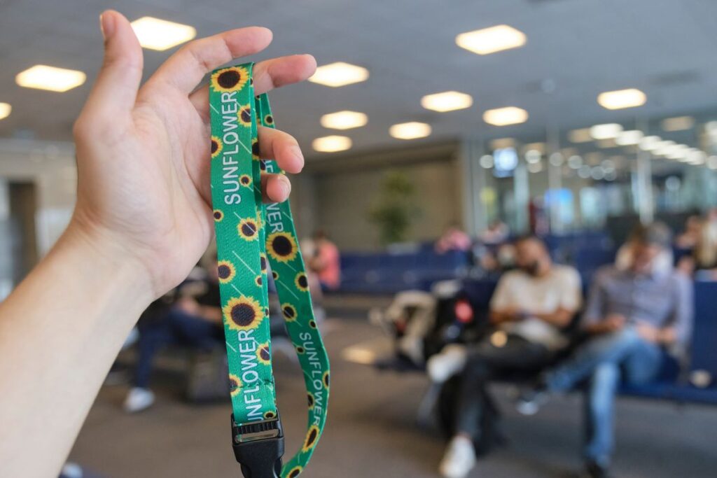 Mão de homem jovem e branco levantada mostrando o cordão de girassol (um dos Cordões de Acessibilidade), por de trás desfocado há um ambiente de sala de espera de um aeroporto.