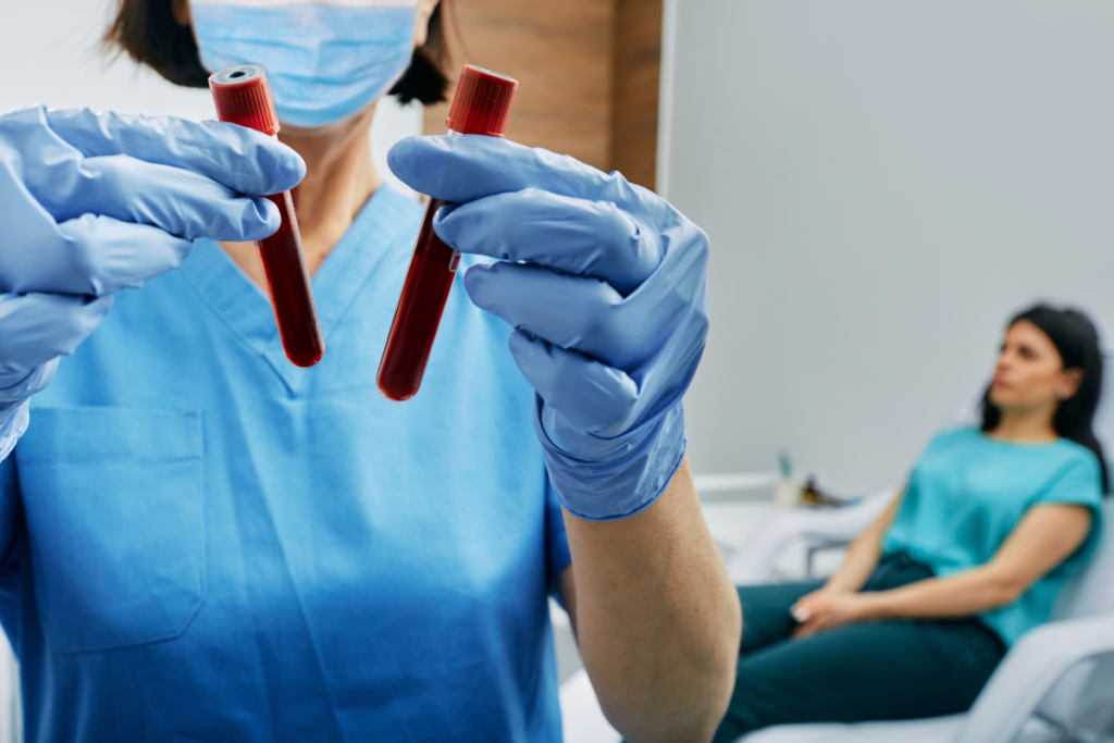 A foto ilustra um momento de coleta de exame de sangue, efetivo para diagnosticar Linfoma ou outras doenças do sangue. Foto de uma profissional de laboratório, com dois tubos de sangue coletados. A paciente ao fundo desfoca, está sentada aguardando liberação.