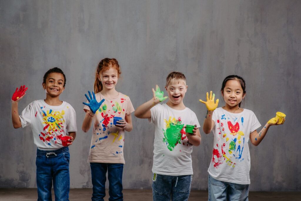 Esta imagem representa a diversidade e a inclusão na sua forma mais pura, com quatro crianças felizes e sorrindo. Todas estão usando blusas pintadas com tinta e mostrando as mãos abertas sujas de tintas coloridas. As crianças possuem características diferentes, sendo um menino negro, uma menina ruiva, um menino loiro com Síndrome de Down e uma menina asiática.